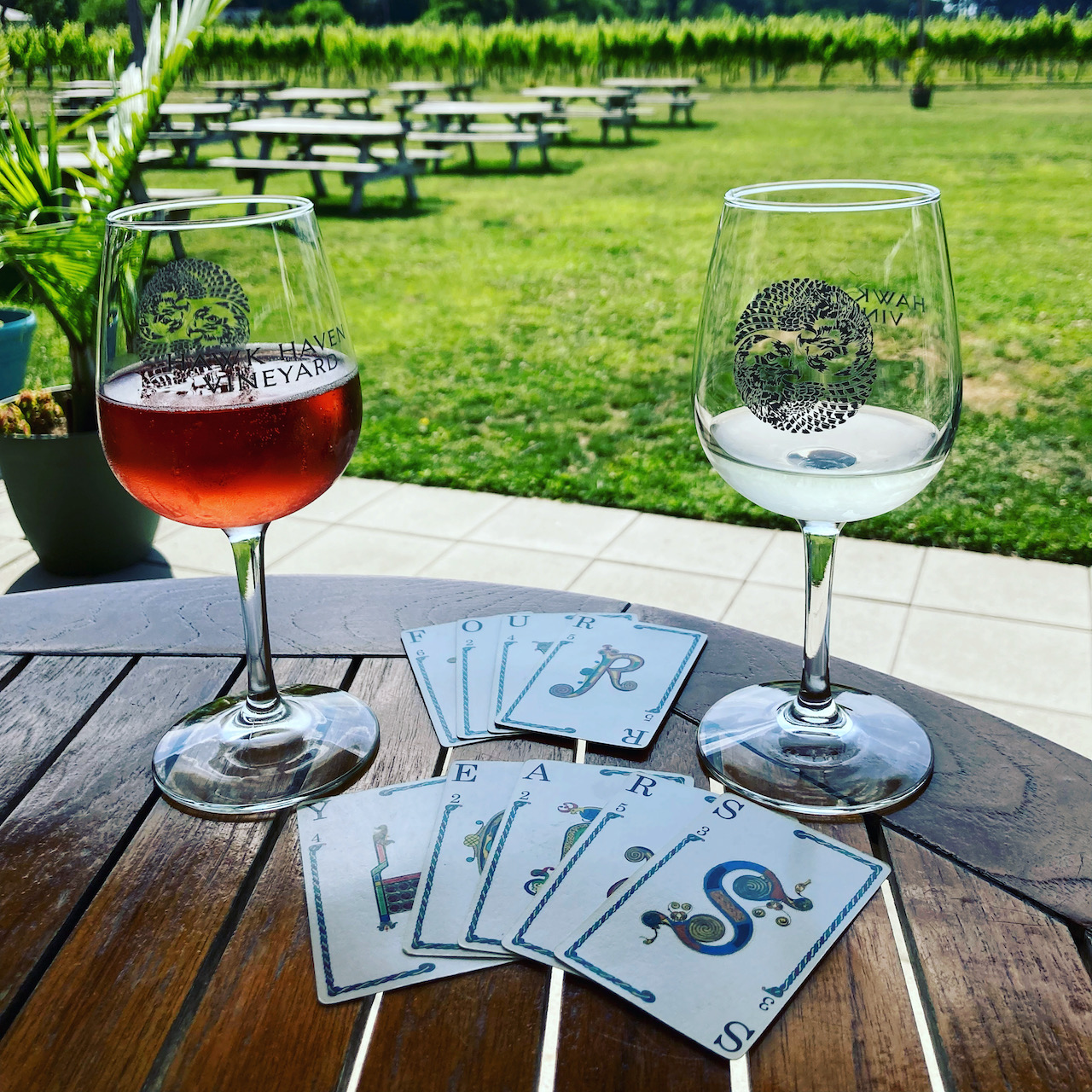 Two wine glasses on wooden table with vineyard in background. On table in foreground are playing cards that spell out FOUR YEARS.