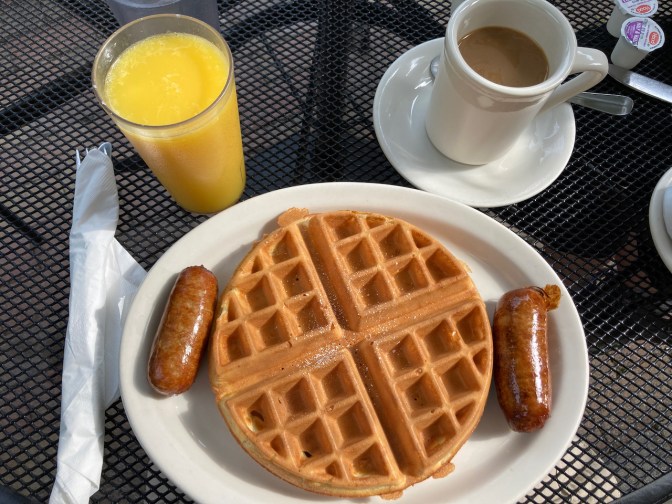 Belgian waffle with two sausages on white plate, with glass of orange juice and mug of coffee on picnic table.