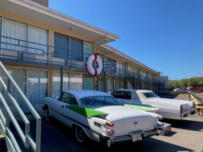 Exterior of Lorraine Motel, with two 1960s cars parked in front of building and wreath on railing outside Room 306.