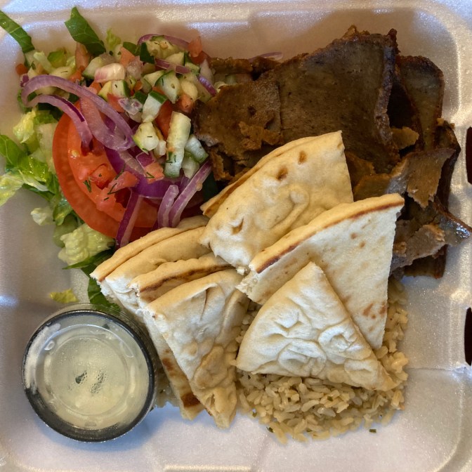 Gyro platter with pita bread, salad, rice, and sauce in a styrofoam container.