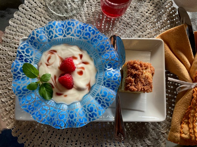 Yoghurt in blue bowl, and coffee cake on white plate, on table.
