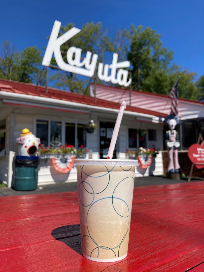 Milk shake in cup on table with exterior of Kayla Drive-In in background.