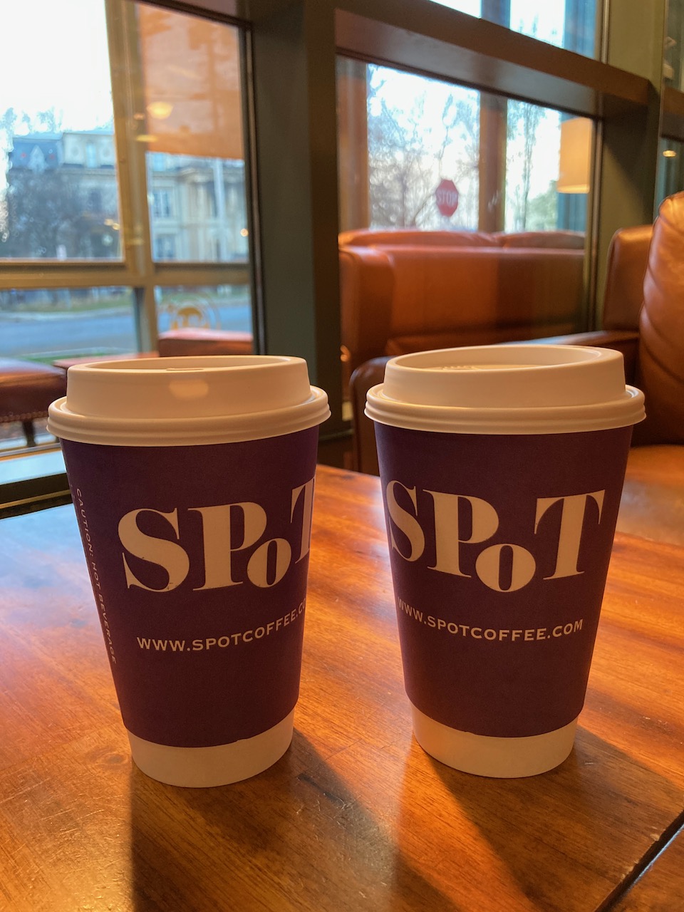 Two cups of coffee that say SPOT, on a wooden table in front of a window.