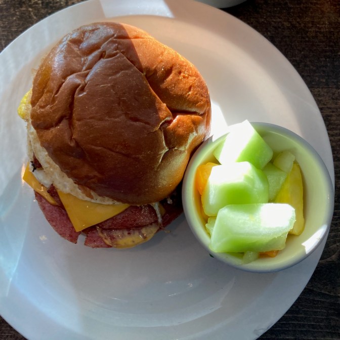 White plate with small bowl of fruit, along with breakfast sandwich.