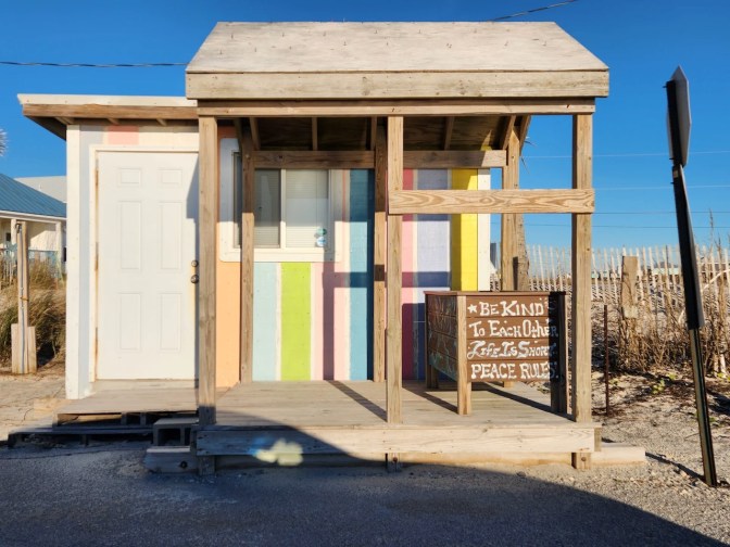 Small beachfront shack with signs on porch that reads BE KIND OF EACH OTHER LIFE IS SHORT PEACE RULES.