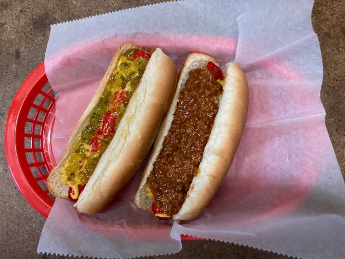 Two hot dogs, one covered in relish and mustard, the other covered in chili.