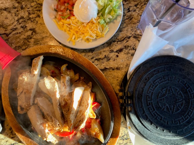 Chicken fajita on sizzling platter, with plate that has cheese, lettuce, and salsa, and container of flour tortillas.