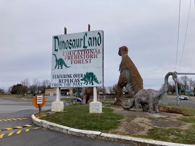 Sign that reads DINOSAUR LAND EDUCATIONAL HISTORIC FOREST beside scale models of dinosaurs.