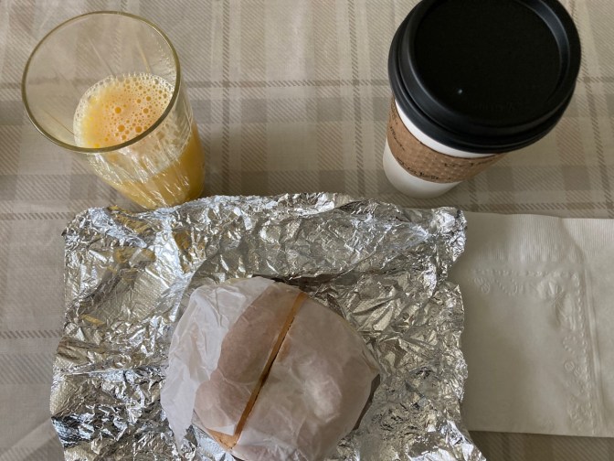 Bagel sandwich on table, wrapped in tin foil, with cup of orange juice and cup of coffee.