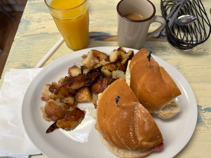 Pork roll, egg, and cheese sandwich on kaiser roll on plate, with side of potatoes. A cup of coffee and a glass of orange juice are also on table.
