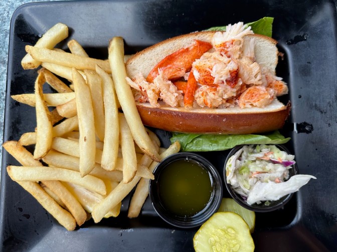 Lobster roll, fries, and coleslaw on black plate.