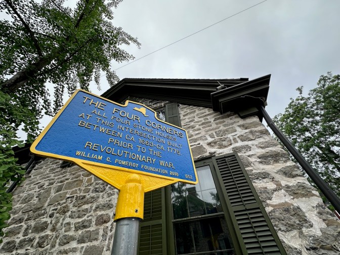 Blue sign that says THE FOUR CORNERS ALL FOUR STONE HOUSES AT THIS INTERSECTION BUILT BETWEEN CA 1663-1775 PRIOR TO THE REVOLUTIONARY WAR.