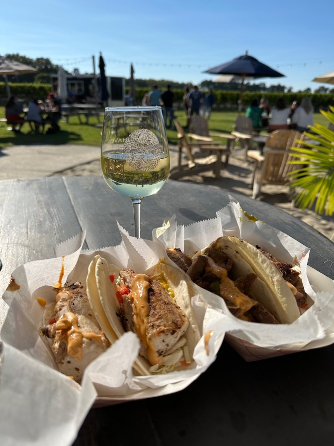 Two dishes of tacos on paper plates, with glass of wine behind them.