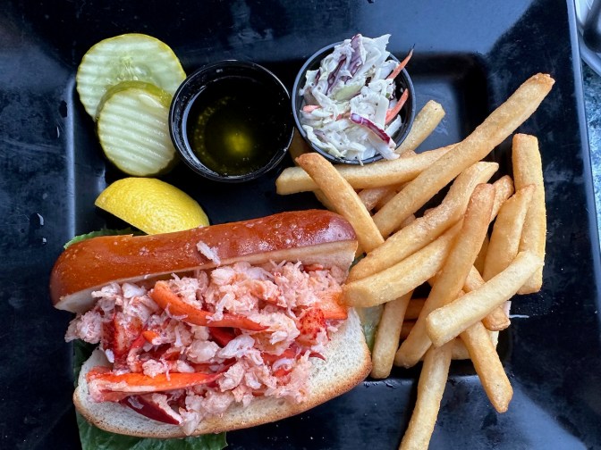 Lobster roll, French fries, coleslaw, pickles, and cup of butter on black plate.