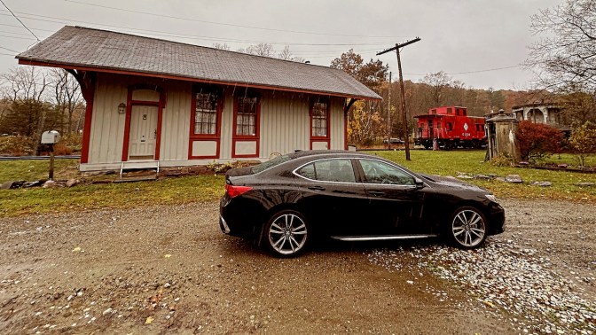 Acura TLX, parked beside old train station. A caboose is in the background.
