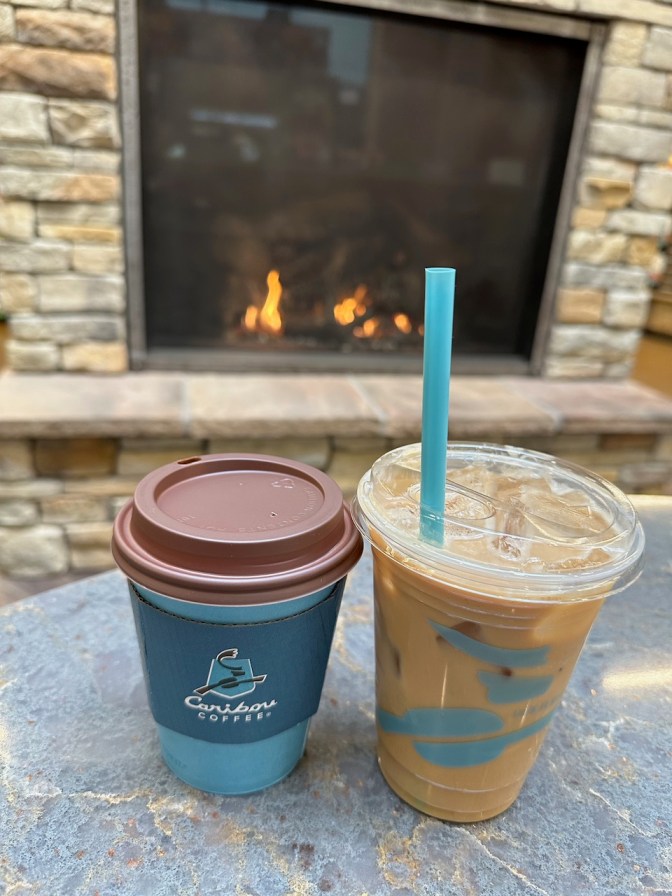 Hot coffee in paper cup and iced oatmilk latte in clear plastic cup on table near fireplace.