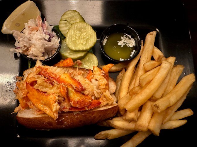Lobster roll on plate, with French fries, coleslaw, and pickles.
