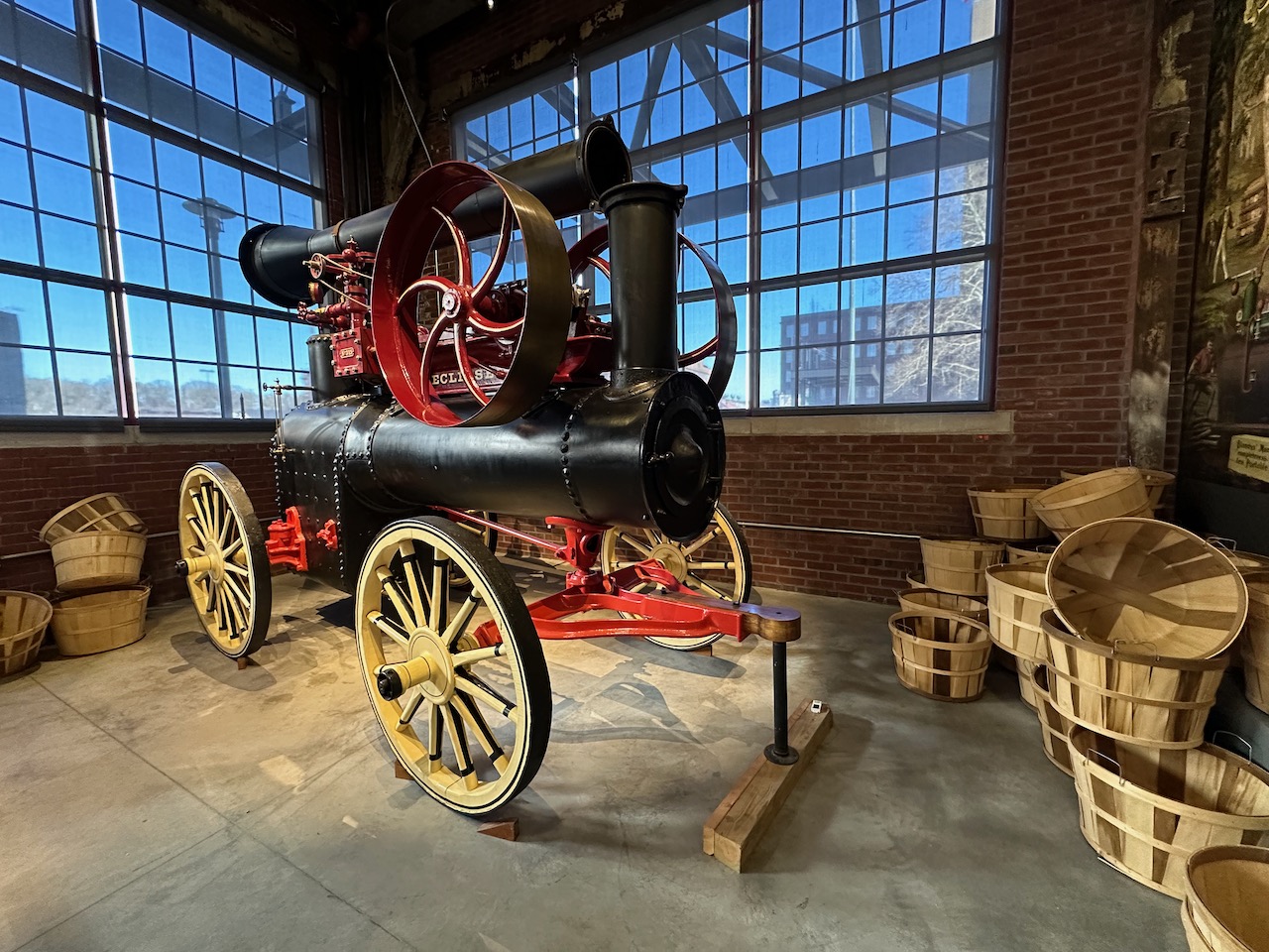 Portable steam engine in corner of building. Brick walls and glass windows are behind the machine.