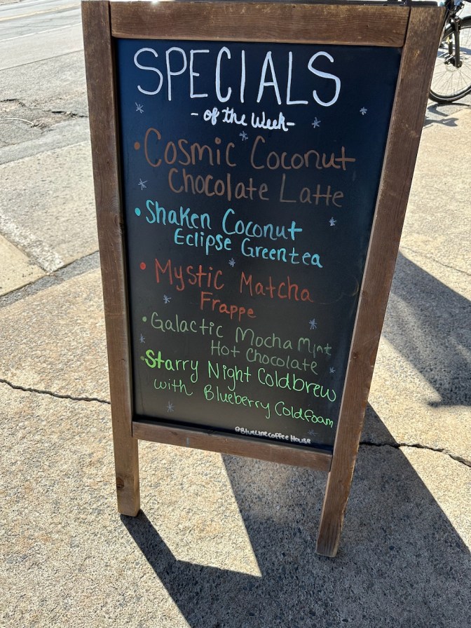 Chalkboard menu outside Blue Line Coffee House listing special including COSMIC COCONUT CHOCOLATE LATTE, SHAKEN COCONUT ECLIPSE GREEN TEA, MYSTIC MATCHA FRAPPE, GALACTIC MOCHA MINT HOT CHOCOLATE, AND STARRY NIGHT COLDBREW WITH BLUEBERRY COLDFOAM.