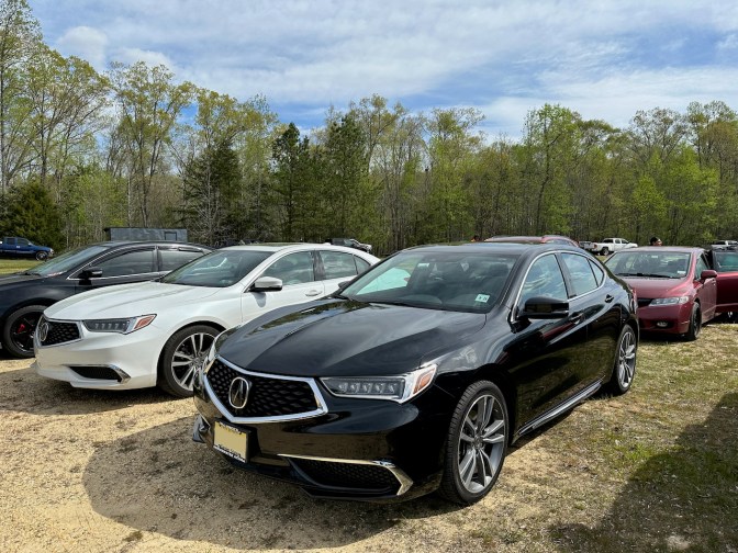 2020 Acura TLX in black parked beside a white Acura TLX in grass and dirt parking lot.