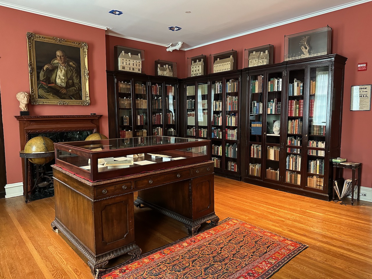 Large room with floor-to-ceiling book cases and a large display case in middle of room. A portrait hangs over a fireplace on the left side of the image.