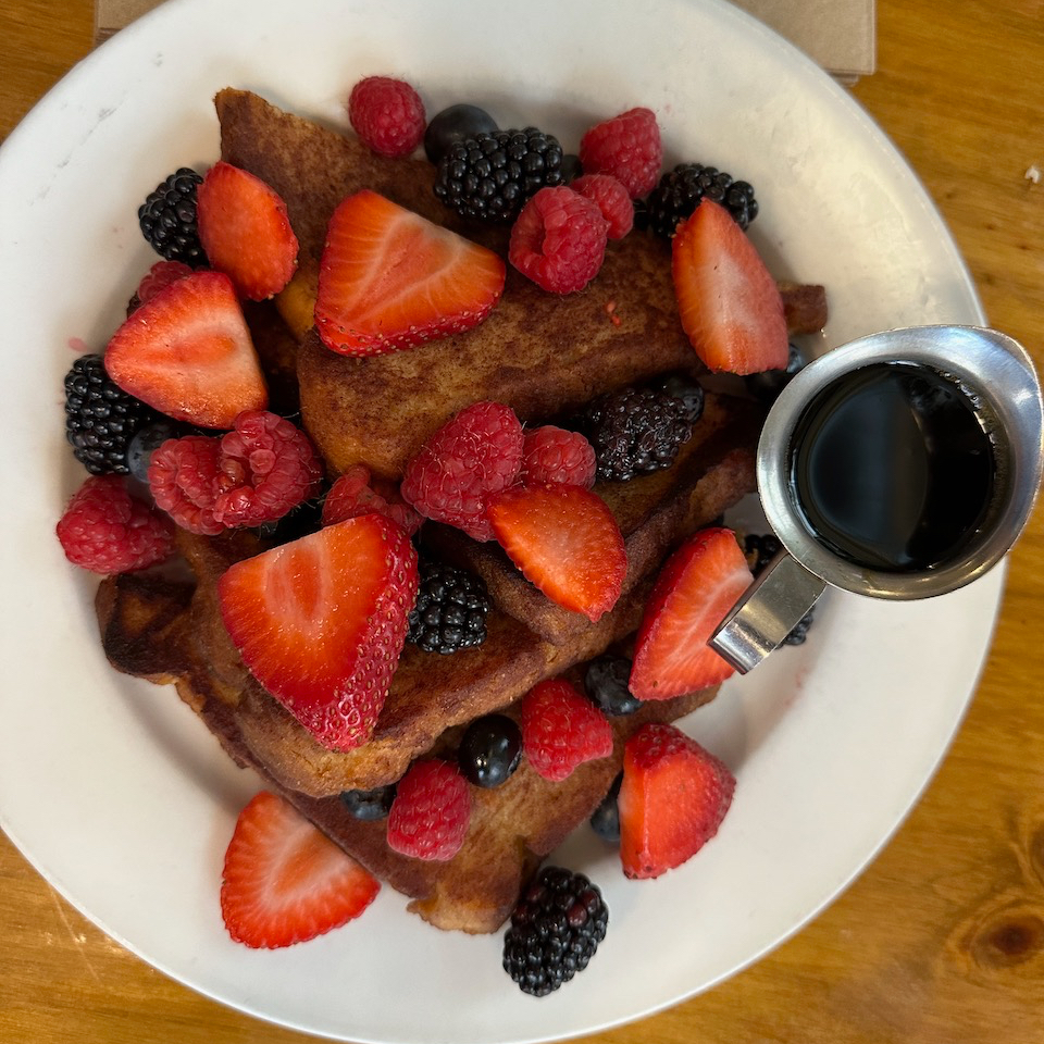 French toast, topped with mixed fruit, on a white plate. A small pitcher of maple syrup sits to the side.