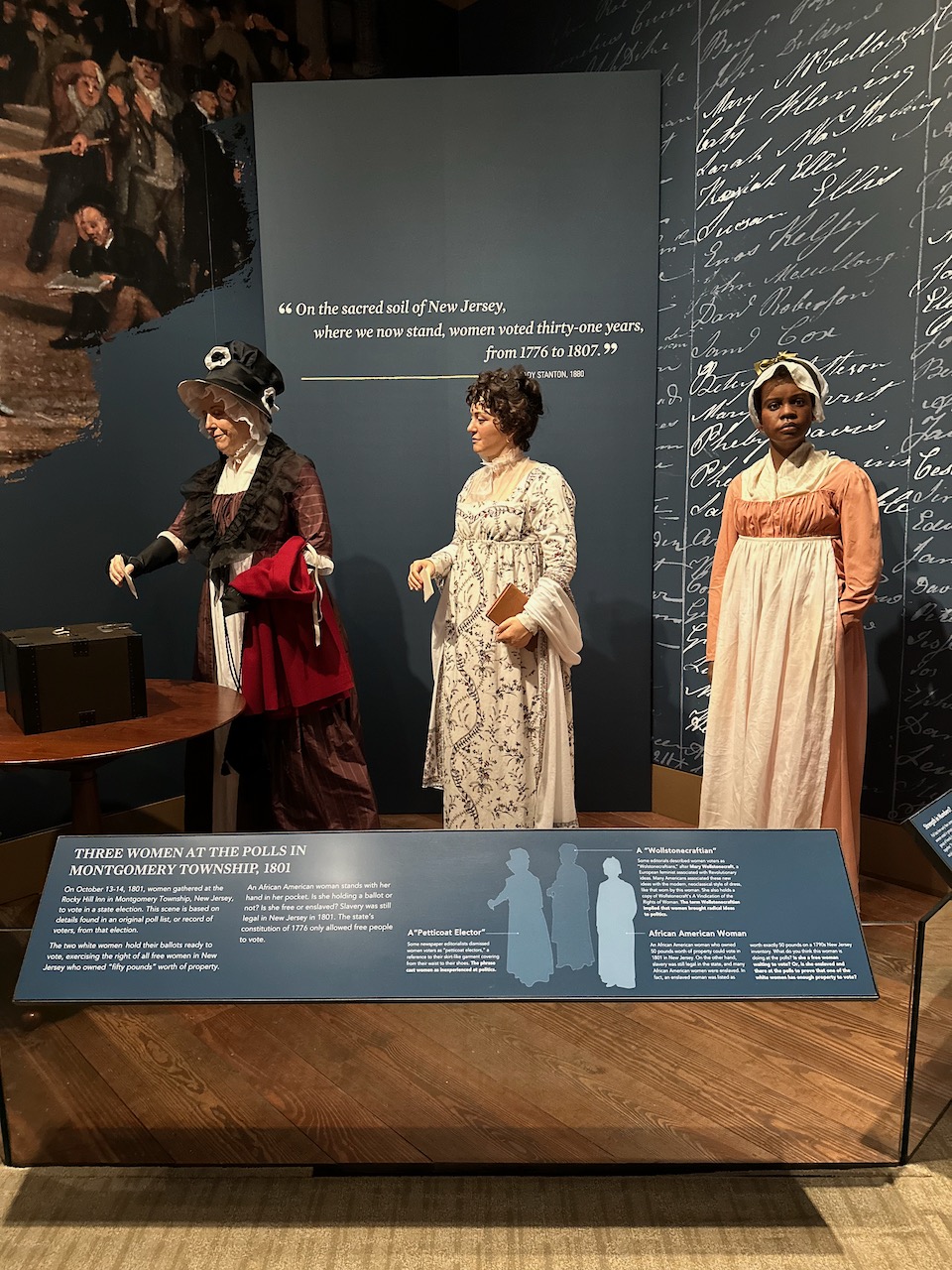 Display of three women in line to vote at a polling station. The mannequins are dressed in late 18th century outfits.