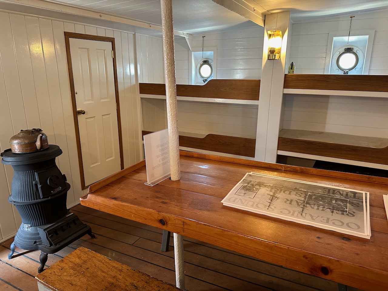 Crew quarters, with wooden table in middle of room, bunks along walls, and pot belly stove near entrance.
