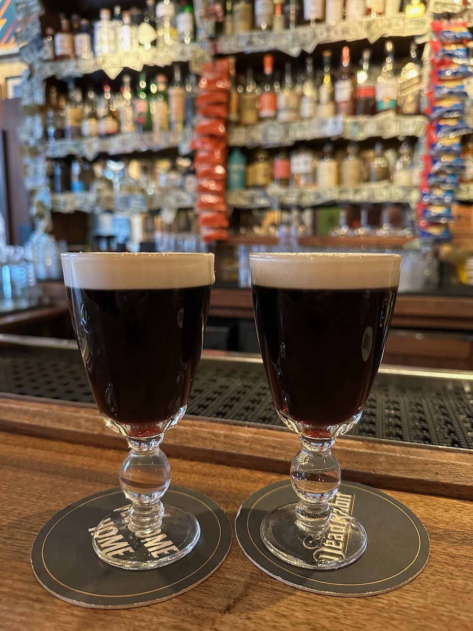 Two Irish coffees in glasses. The coffee glasses are on the bar, on coasters.