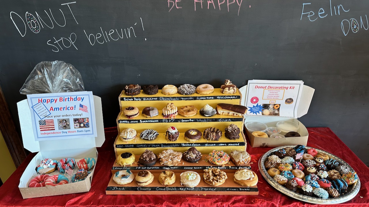 Table with assortment of 30 different donuts on display, along with gift boxes of donuts. A blackboard with writing is in background.