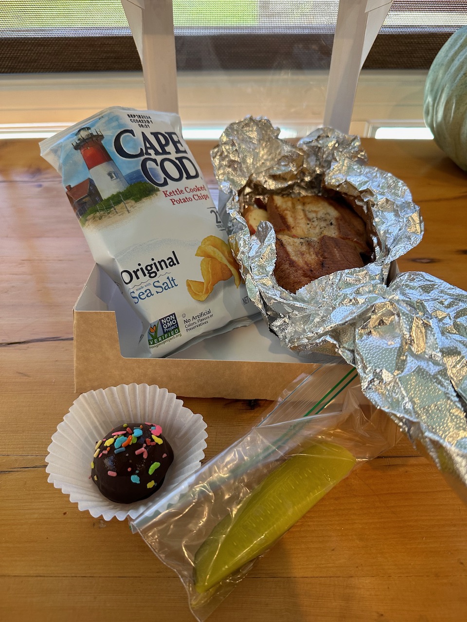 Countertop with box including sandwich, potato chips, pickle in plastic bag, and small cookie.