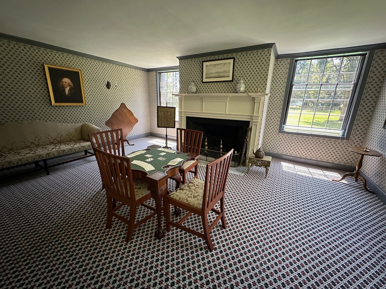 Parlor room of Schuyler House, with framed painting of George Washington hanging on wall and playing card table with four chairs in center of room. A fireplace is on the far side wall.