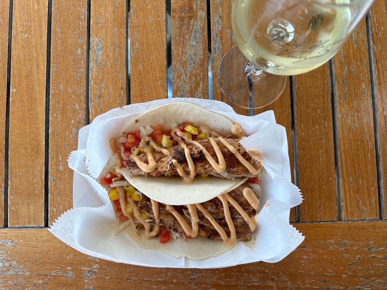 Two fish tacos in paper bowl on wooden table. A glass of wine is nearby.