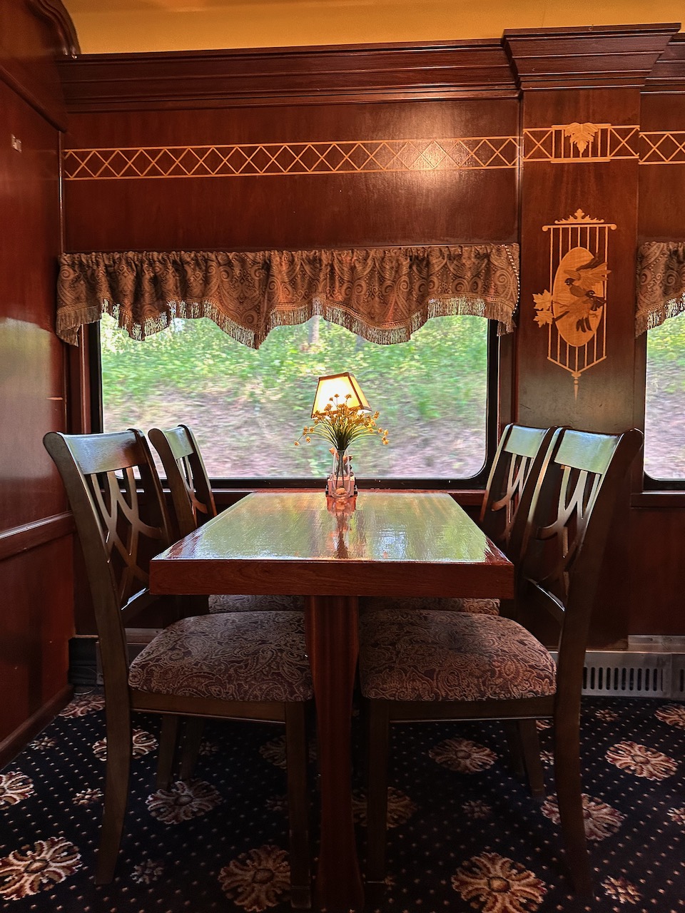 Table with four wooden chairs in dining car. There is a blue and white carpet on the floor, and a window behind the table looks out at trees.