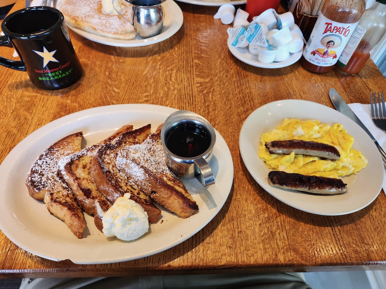 French toast, eggs, and sausage on white plates on wooden table.