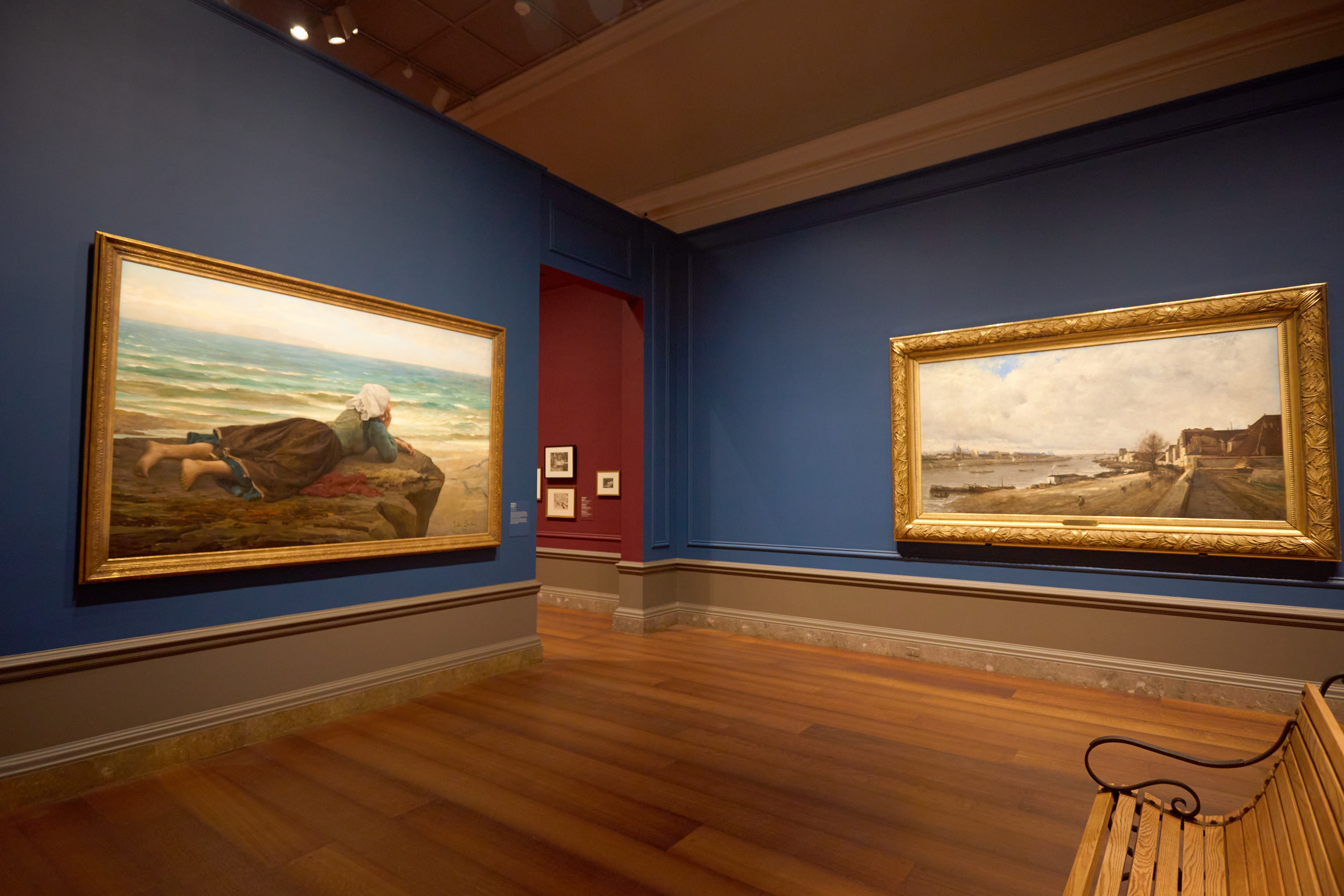 Two paintings on display in gallery. On left a girl sits on the beach looking at the ocean. On the right is a landscape of a town beside a canal.
