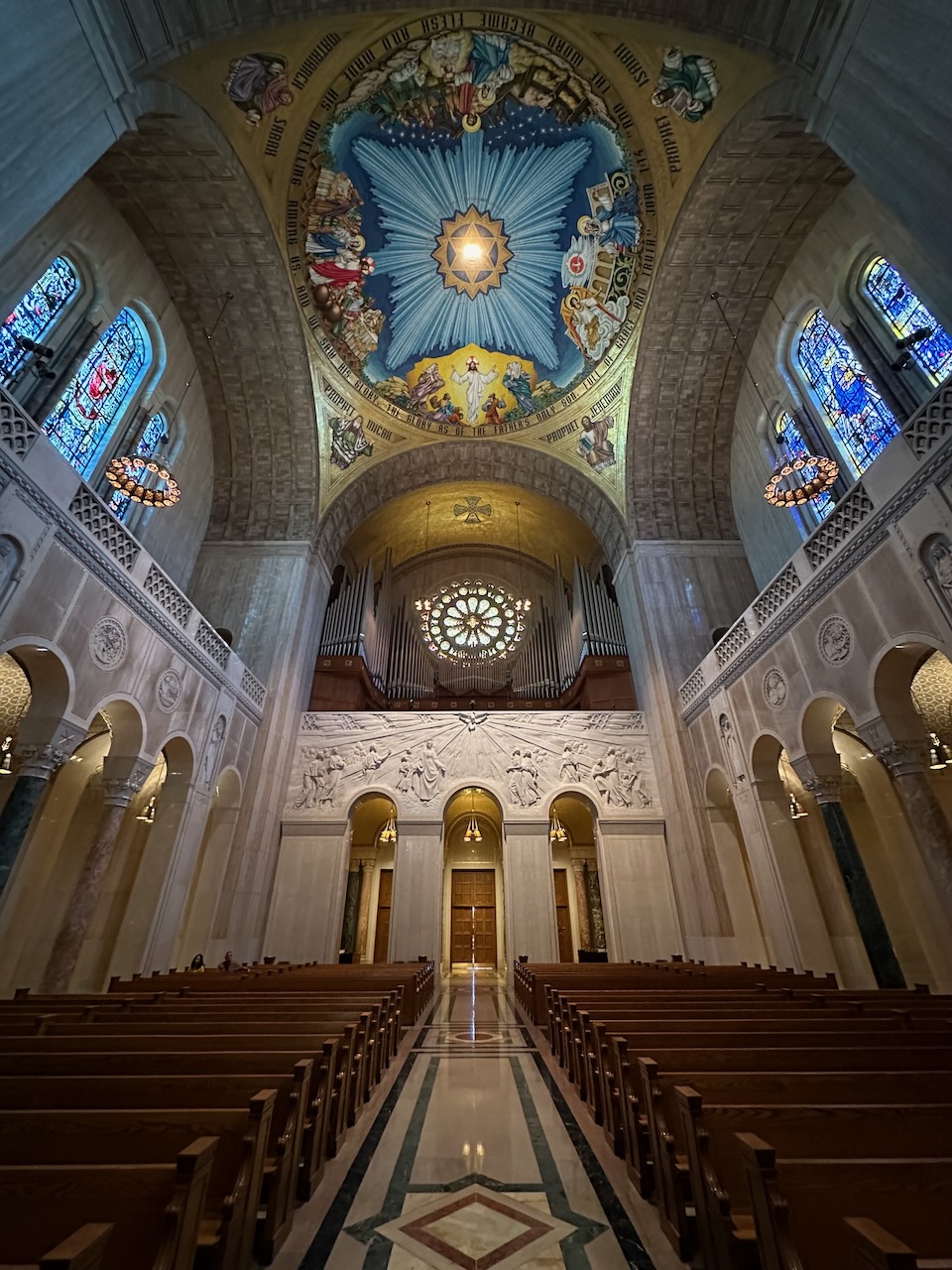 Interior of basilica, with pews, stained glass windows, and mosaics on ceiling.