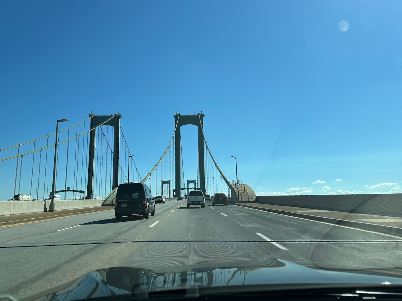 View of Delaware Memorial Bridge from behind dashboard of car. The sky is blue with only a few clouds on the horizon.