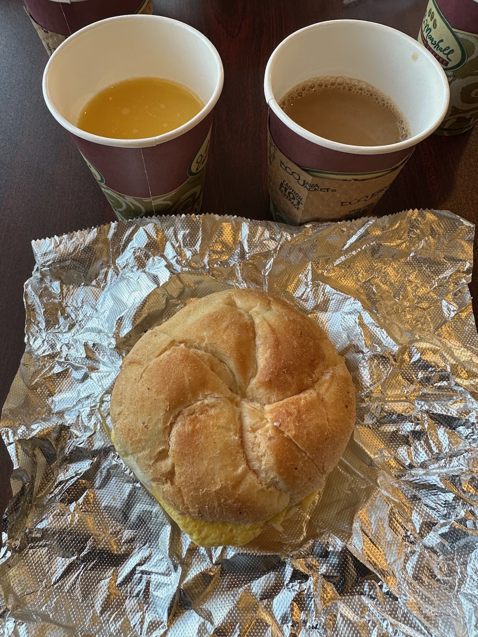 Breakfast sandwich on foil wrapper, with cup of coffee and cup of orange juice in background.