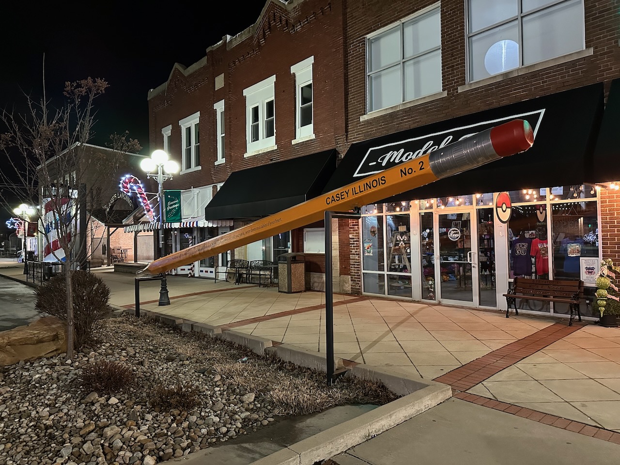 Large pencil outside of buildings along Main Street. The Pencil is supported by two metal poles.