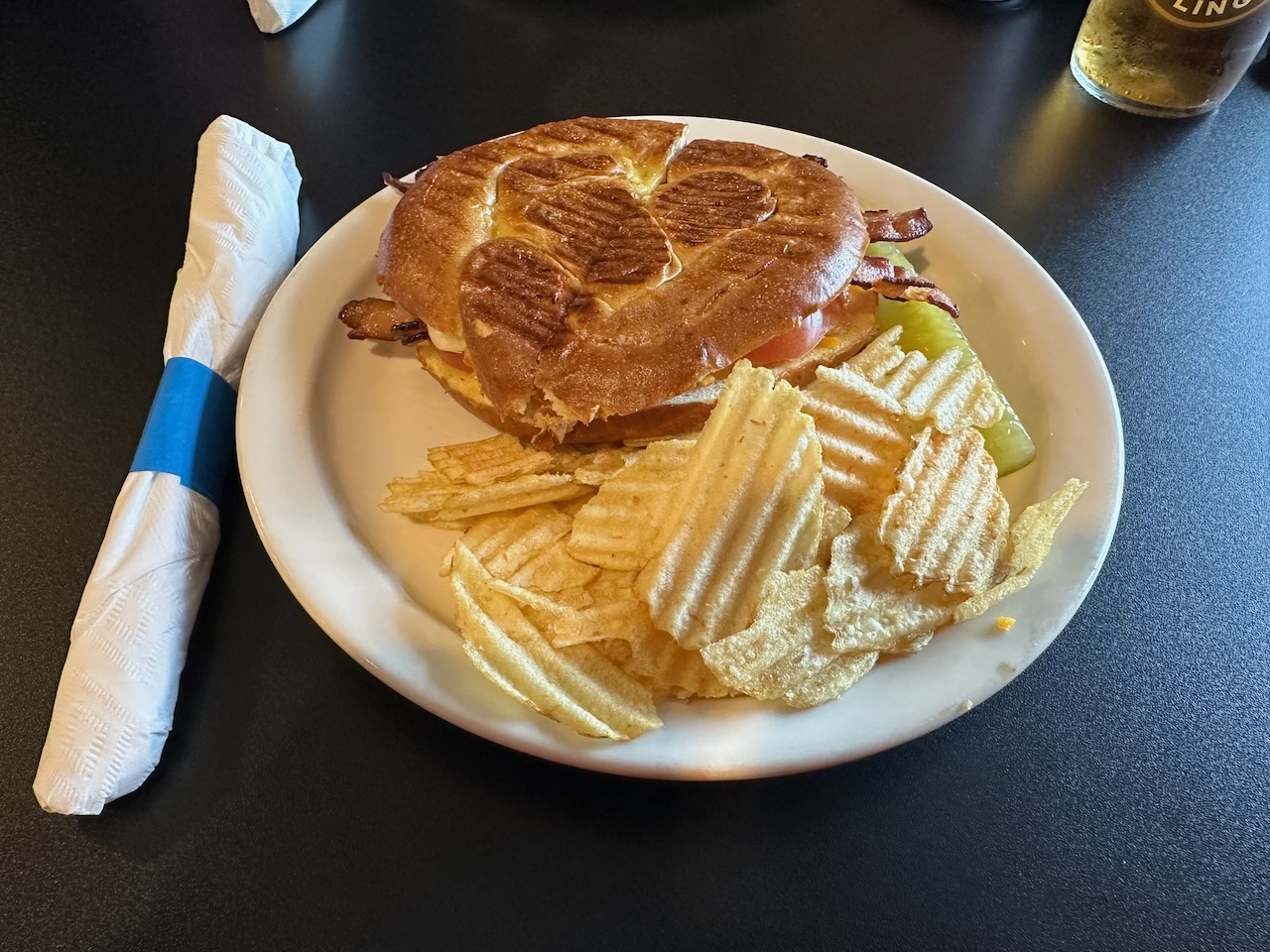 Sandwich on pretzel roll with side of potato chips and pickle, on white plate.