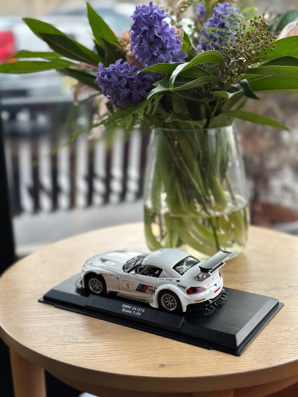 Small model of BMW racer on stand. The car is on a wooden table with a vase of flowers behind it. 