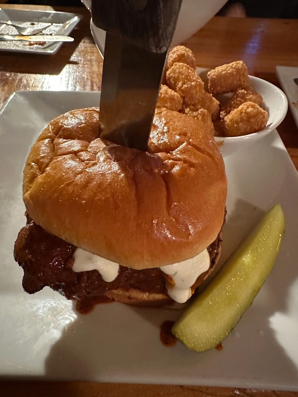 Chicken sandwich on plate with knife in it, and a side of tater tos. 