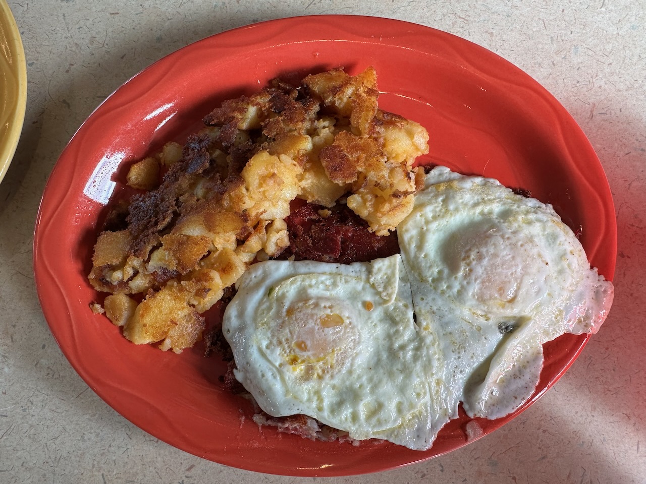 Corned been hash and home fries with eggs on red plate. 