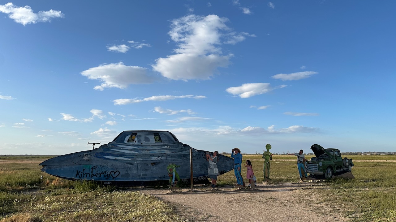 Statue of UFO and car with statues of people and aliens on side of road.