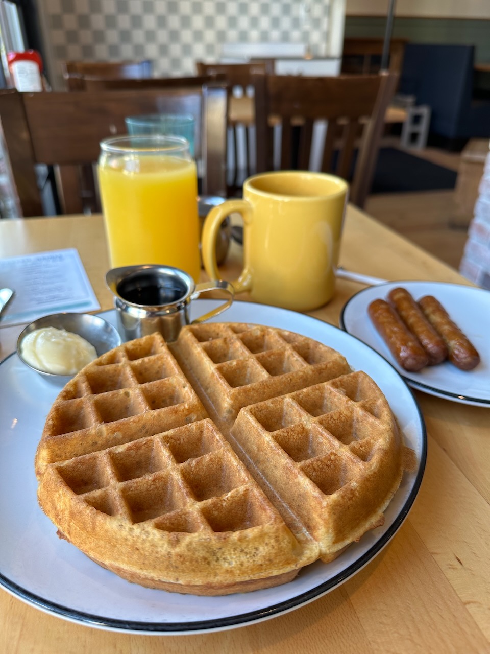 Plate with waffle, beside smaller plate with sausage links, glass of orange juice, and coffee cup on table. 