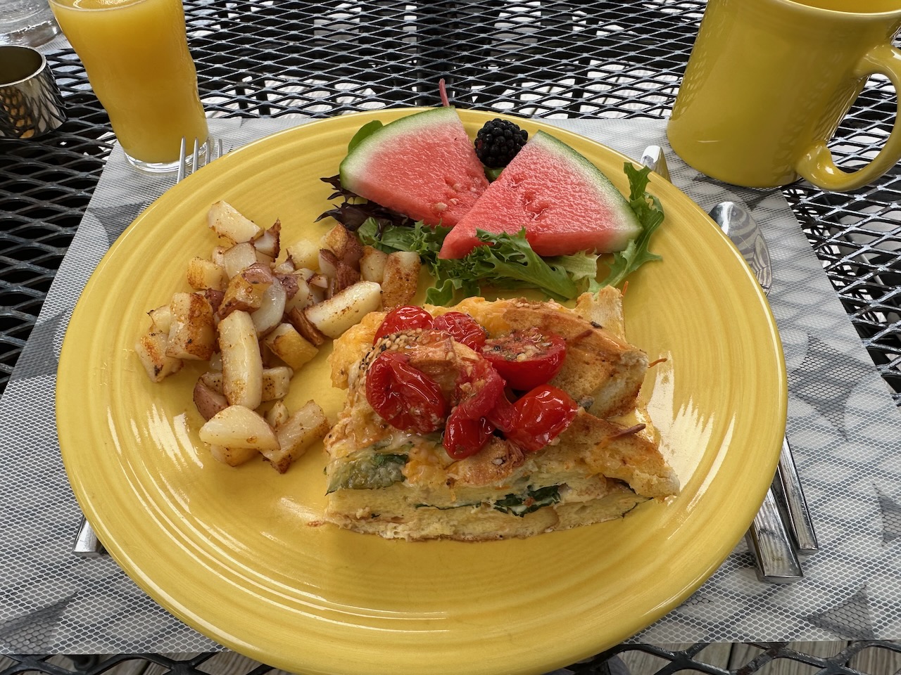 Yellow plate with potatoes, watermelon, and a bagel and egg bread pudding. 