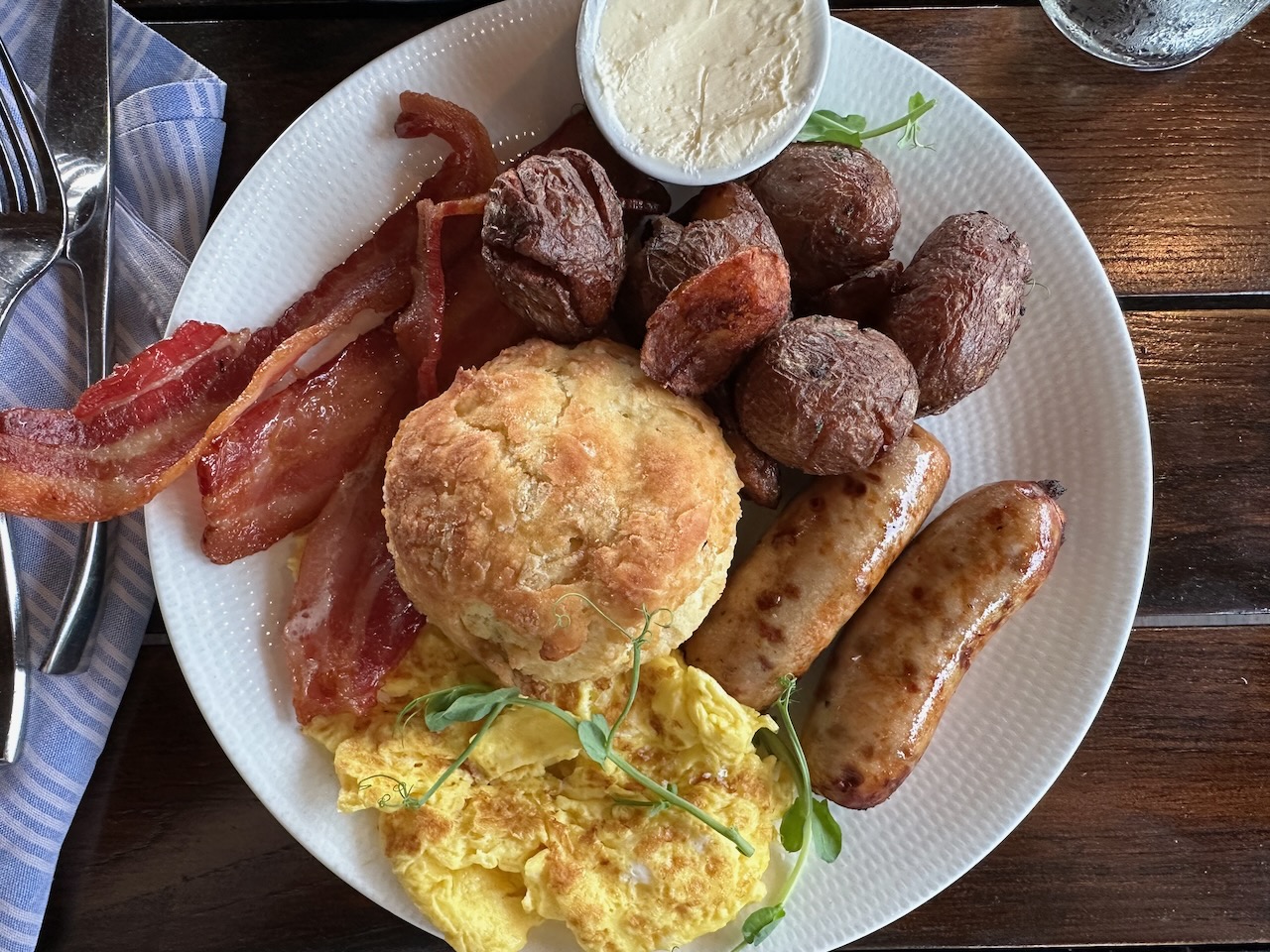 White plate with eggs, sausage, bacon, potatoes, and biscuit. 