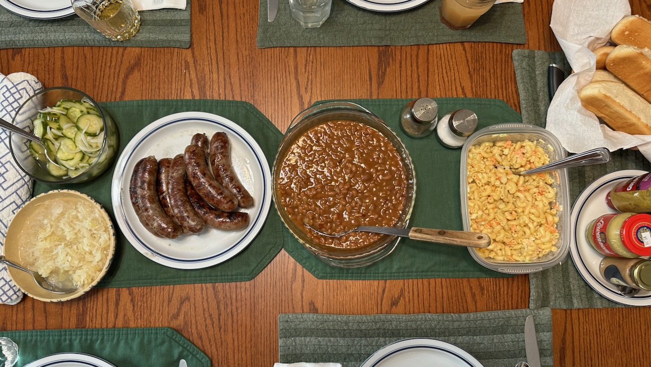 Table with bratwurst, baked beans, macaroni salad, and sides.