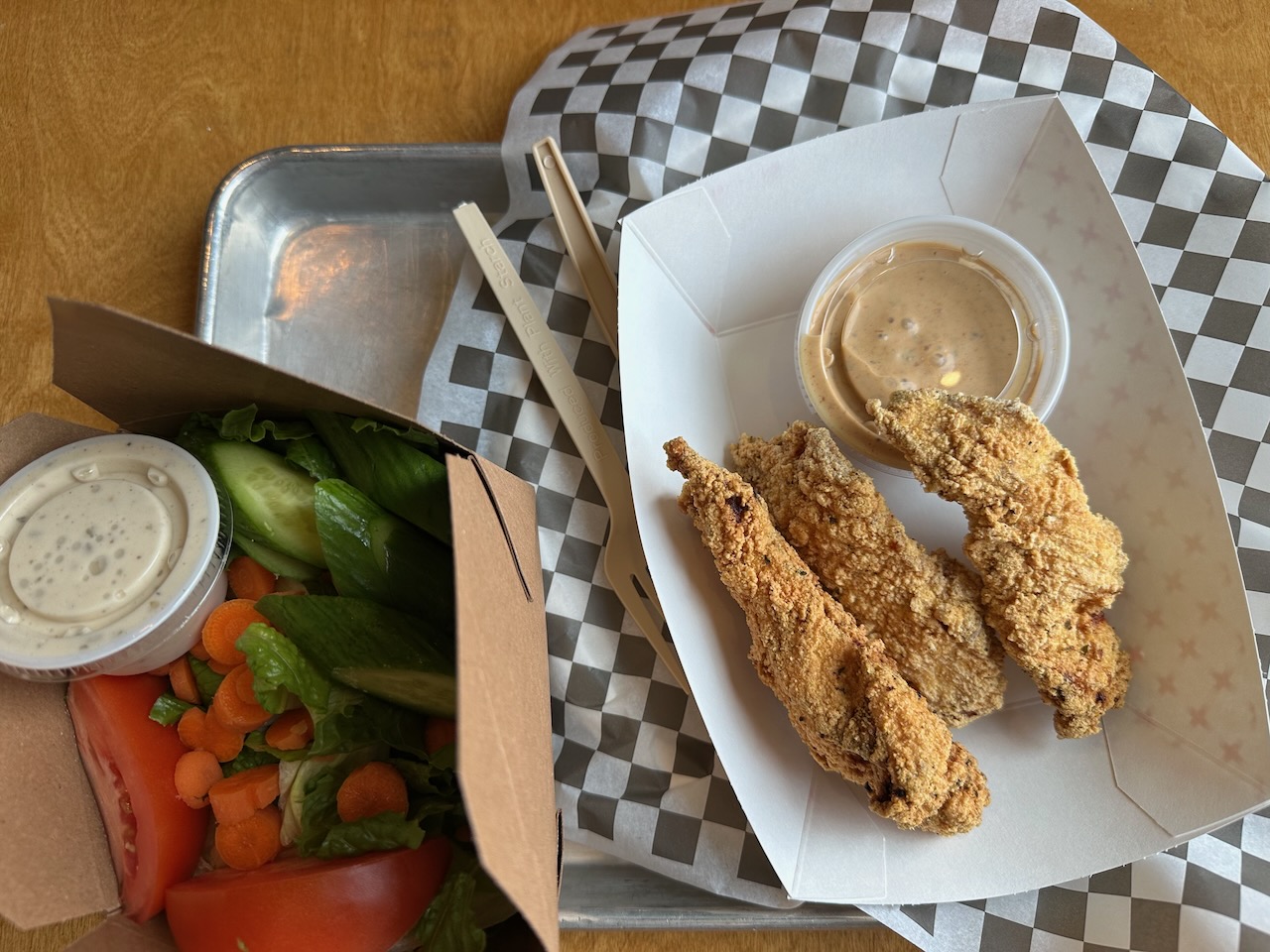 Chicken fingers in white paper dish and basket with salad, on a wooden table.